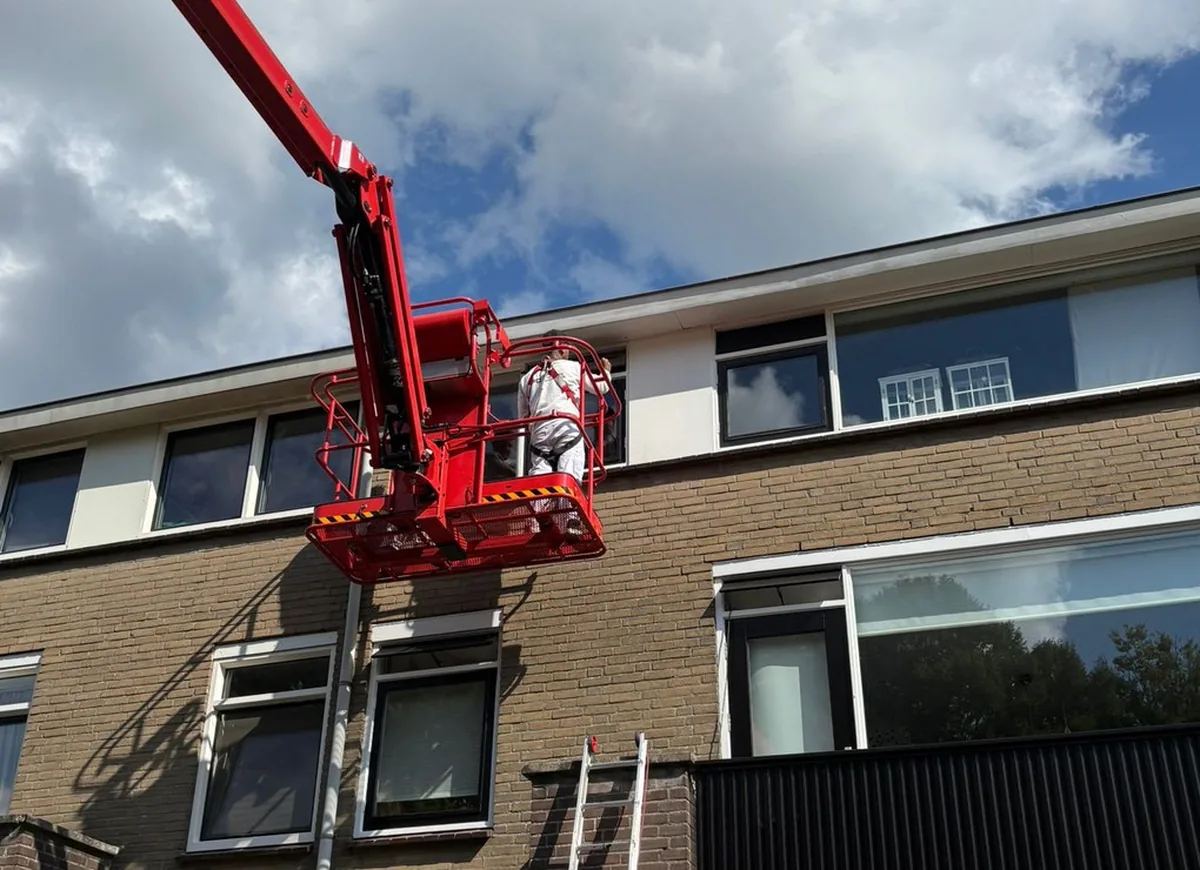Een medewerker in de bak van een rode hoogwerker werkt aan de gevel van een woonblok